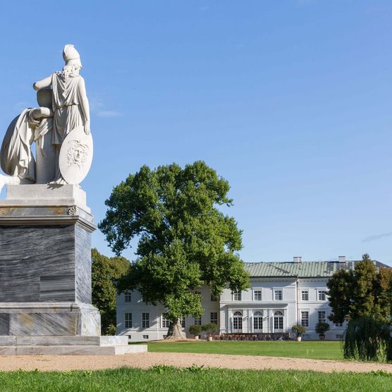 Eine steinerne Skulptur steht im Schlosspark Neuhardenberg und im Hintergrund zeichnet sich das Schloss ab.