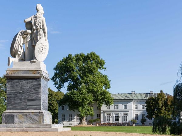 Eine steinerne Skulptur steht im Schlosspark Neuhardenberg und im Hintergrund zeichnet sich das Schloss ab.