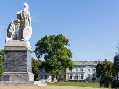 Eine steinerne Skulptur steht im Schlosspark Neuhardenberg und im Hintergrund zeichnet sich das Schloss ab.