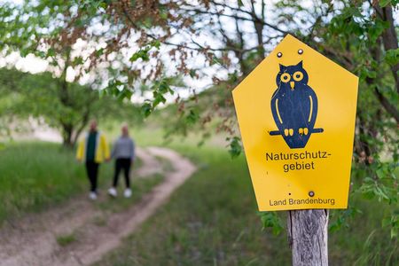 Die schwarze Naturschutzeule auf einem gelben Schild weißt ein Naturschutzgebiet in Brandenburg aus. Im Hintergrund sind zwei Wanderer zu sehen.