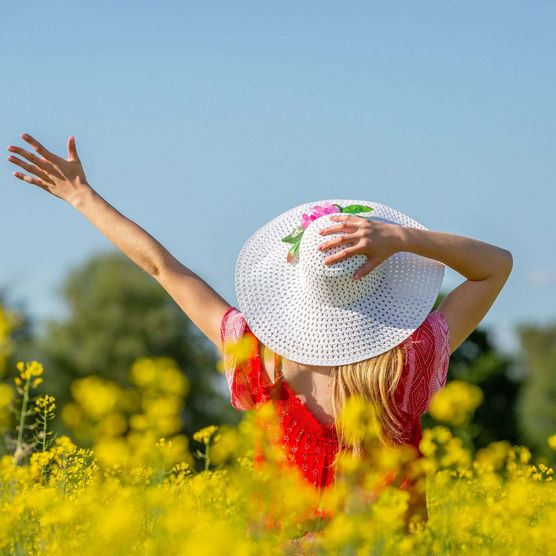 Eine junge Frau mit Sommerhut steht inmitten eines Rapsfeldes