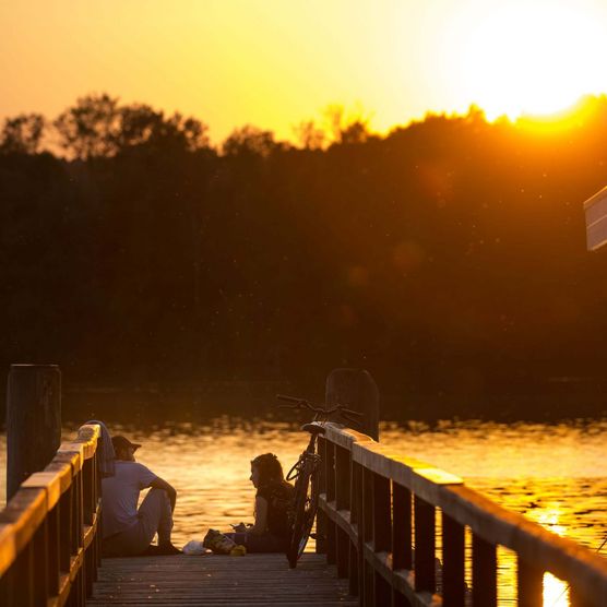 Freunde sitzen während des Sonnenuntergangs auf einem Steg am Scharmützelsee in der Nähe vom Cecilienpark und einem Hausboot am Steg.