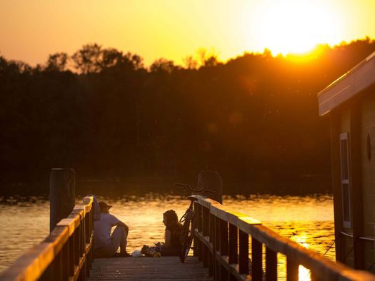 Freunde sitzen während des Sonnenuntergangs auf einem Steg am Scharmützelsee in der Nähe vom Cecilienpark und einem Hausboot am Steg.