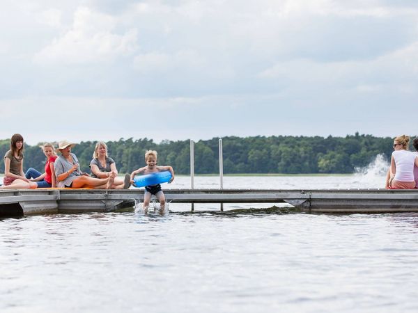 Ein Junge paddelt mit blauem Schwimmring und wird von einem älteren Mädchen schwimmend begleitet. 