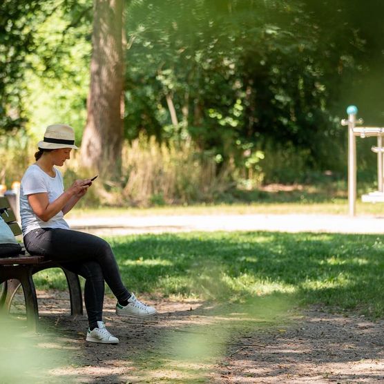 Eine junge Frau sitzt auf einer Bank im grünen Schlosspark Schöneiche bei Berlin und informiert sich in ihrem Handy