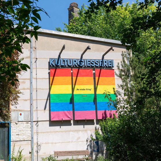 Blick auf die Kulturgießerei Schöneiche bei Berlin, an dessen Wand ein Schild mit der Aufschrift "Kulturgiesserei" hängt, sowie 3 Regenbogenflaggen mit der Aufschrift "Nie wieder Krieg".